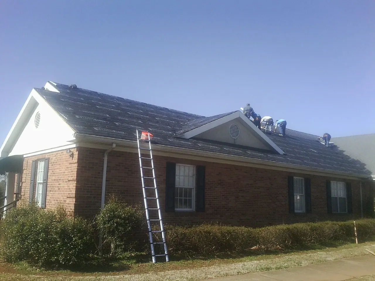Crew tearing shingles off a brick ranch roof with a ladder set — Metal Roof Repair work by our crew in Fowler