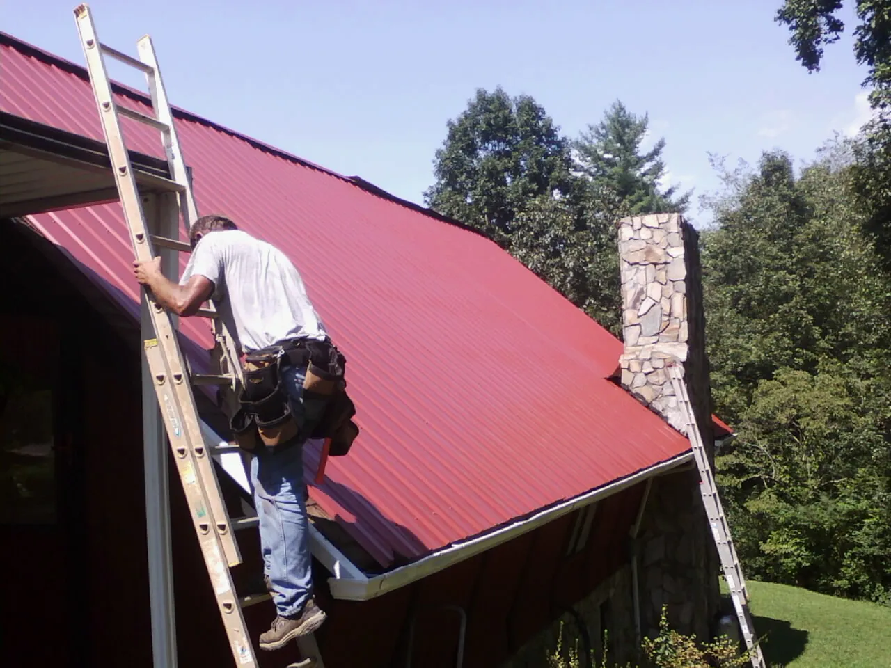Roofer on a ladder beside a red metal roof and stone chimney — Soffit & Fascia Repair by Roofing Brothers in Fowler